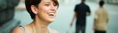 This photo shows a cropped close-up of Cassandra's face. She is laughing and smiling, she has dimples and cropped brown hair, and is wearing a grey tank top and silver necklace. Two figures, out of focus, are walking down a city street in the background. Photo credit: Renik Shchetinin.