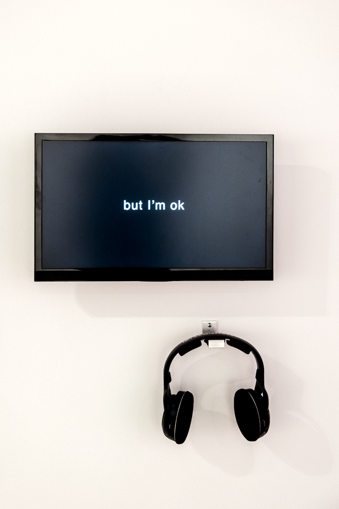 A TV screen, white text on black: "but I'm ok" mounted on a white wall above a pair of over-the-ear headphones.