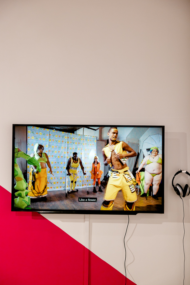 TV showing six people with bodies of different sizes, with black, white, and brown skin, dancing wearing neon yellow, orange and green designer outfits, some holding canes. Caption reads 'like a flower'. There are headphones next to the TV and a pink segment painted on the wall behind. 